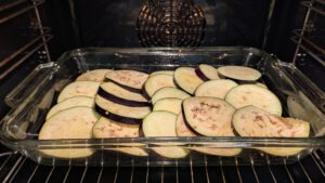 Tranches d'aubergine crues à peau violette, disposées en couche qui se chevauche dans un plat rectangulaire en verre au four ; aucune pomme de terre n'est visible
