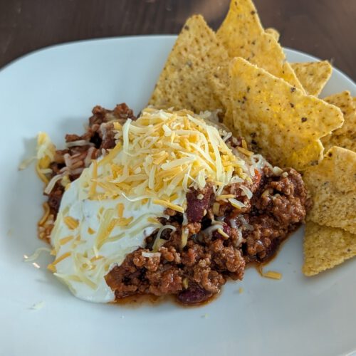 Assiette blanche de chili au bœuf haché et aux haricots rouges, nappé de crème et de cheddar râpé, avec des tortilla chips jaunes à droite, sur une table en bois sombre
