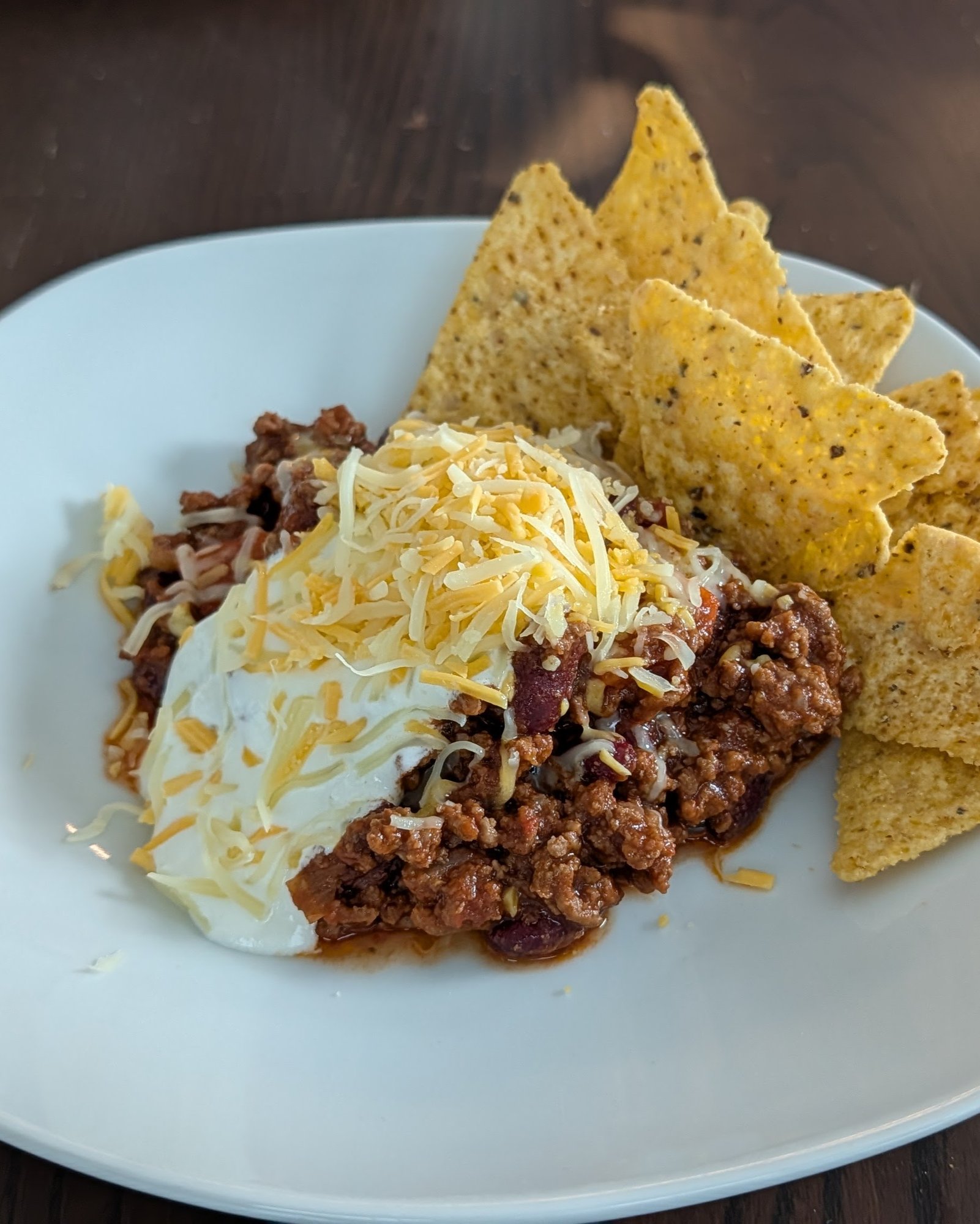 Assiette blanche de chili au bœuf haché et aux haricots rouges, nappé de crème et de cheddar râpé, avec des tortilla chips jaunes à droite, sur une table en bois sombre