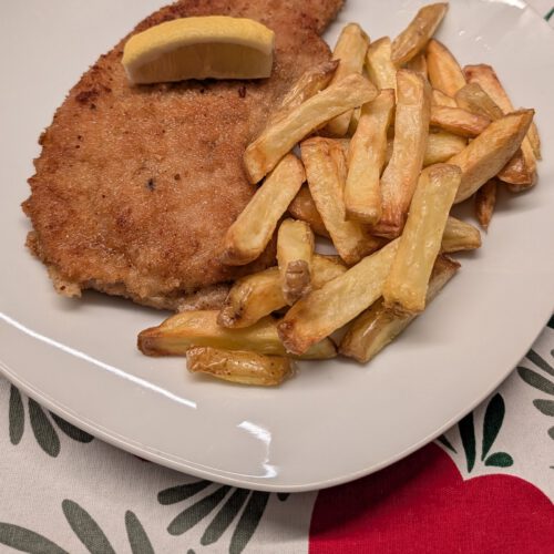 Escalope panée à la croûte dorée dans une assiette blanche, avec un quartier de citron dessus et un tas de frites dorées à côté, sur une nappe à motifs rouge, vert et blanc
