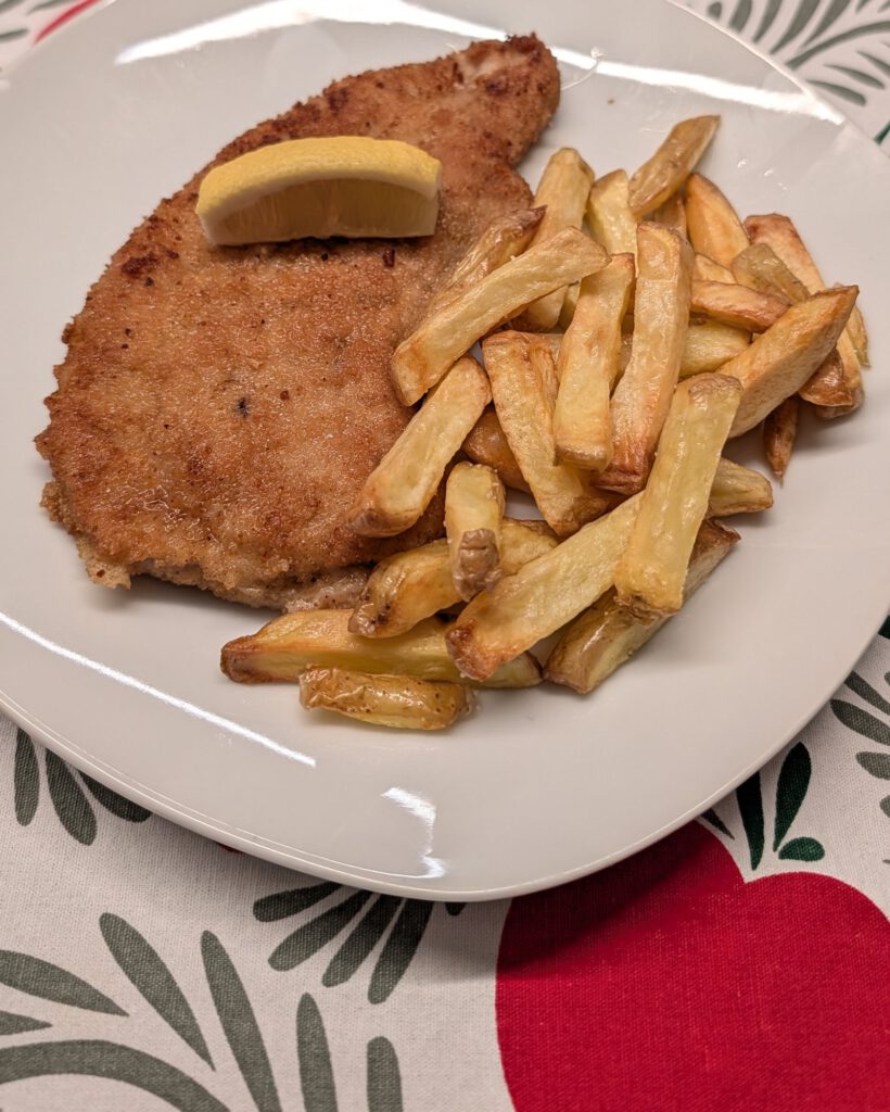 Escalope panée à la croûte dorée dans une assiette blanche, avec un quartier de citron dessus et un tas de frites dorées à côté, sur une nappe à motifs rouge, vert et blanc
