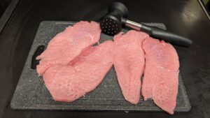 Quatre escalopes crues roses aplaties sur une planche à découper gris foncé, avec un attendrisseur à viande noir posé derrière