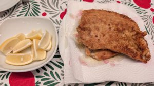Deux escalopes frites et dorées sur du papier absorbant, à côté d'un bol blanc de quartiers de citron, sur une nappe à motifs rouge et vert