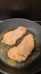 Deux escalopes panées qui cuisent dans une fine couche de graisse, dans une grande poêle sombre, la panure encore pâle sur les bords