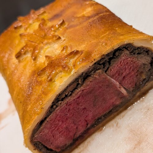Coupe d'un Beef Wellington cuit : bœuf rosé, couche sombre de duxelles et pâte dorée ornée d'un flocon, sur une planche blanche à bord vert