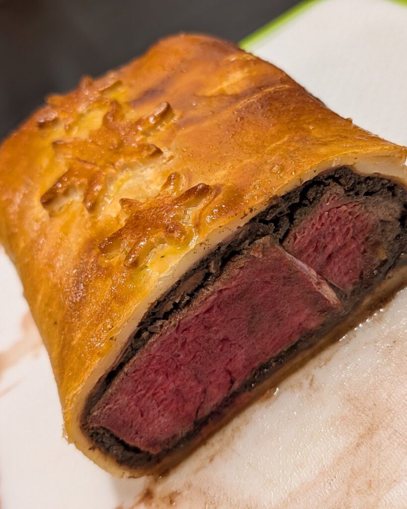 Coupe d'un Beef Wellington cuit : bœuf rosé, couche sombre de duxelles et pâte dorée ornée d'un flocon, sur une planche blanche à bord vert