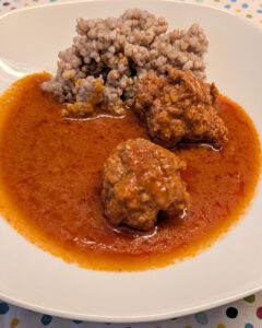 Assiette blanche carrée avec deux boulettes de viande nappées de sauce tomate, un tas de sarrasin cuit et une flaque de sauce orange-rouge sur une nappe à pois