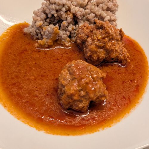 Assiette blanche carrée avec deux boulettes de viande nappées de sauce tomate, un tas de sarrasin cuit et une flaque de sauce orange-rouge sur une nappe à pois