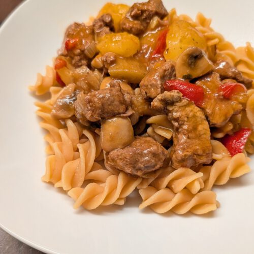 Goulash de bœuf avec morceaux de pommes de terre, champignons et poivron rouge sur des pâtes en spirales, dans une assiette blanche sur une table en bois foncé