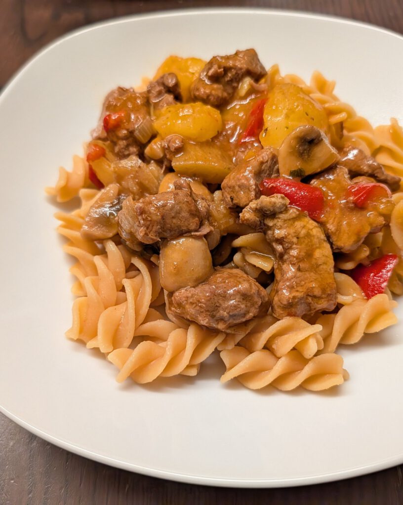 Goulash de bœuf avec morceaux de pommes de terre, champignons et poivron rouge sur des pâtes en spirales, dans une assiette blanche sur une table en bois foncé