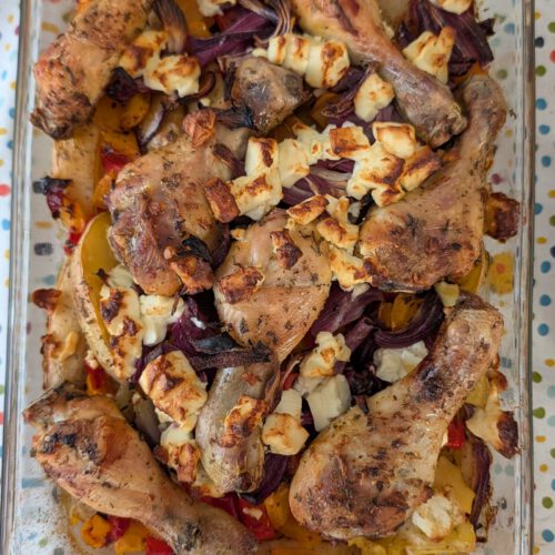 Pilons de poulet dorés dans un plat en verre rectangulaire, avec quartiers de pommes de terre, oignon rouge et poivron, sous des morceaux de feta dorés
