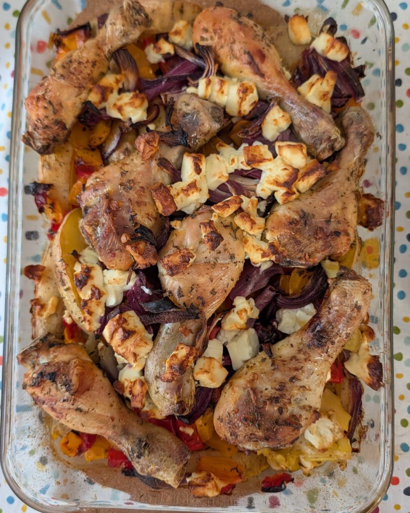 Pilons de poulet dorés dans un plat en verre rectangulaire, avec quartiers de pommes de terre, oignon rouge et poivron, sous des morceaux de feta dorés