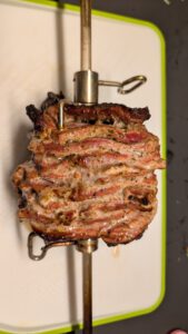 Broche sortie du gril avec le bloc de viande aux couches dorées et croustillantes, posée sur une planche à découper blanche et verte