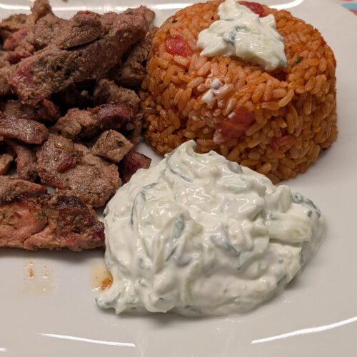 Assiette blanche carrée avec des morceaux de gyros saisis, un dôme de riz à la tomate surmonté de tzatziki et une portion de tzatziki au concombre