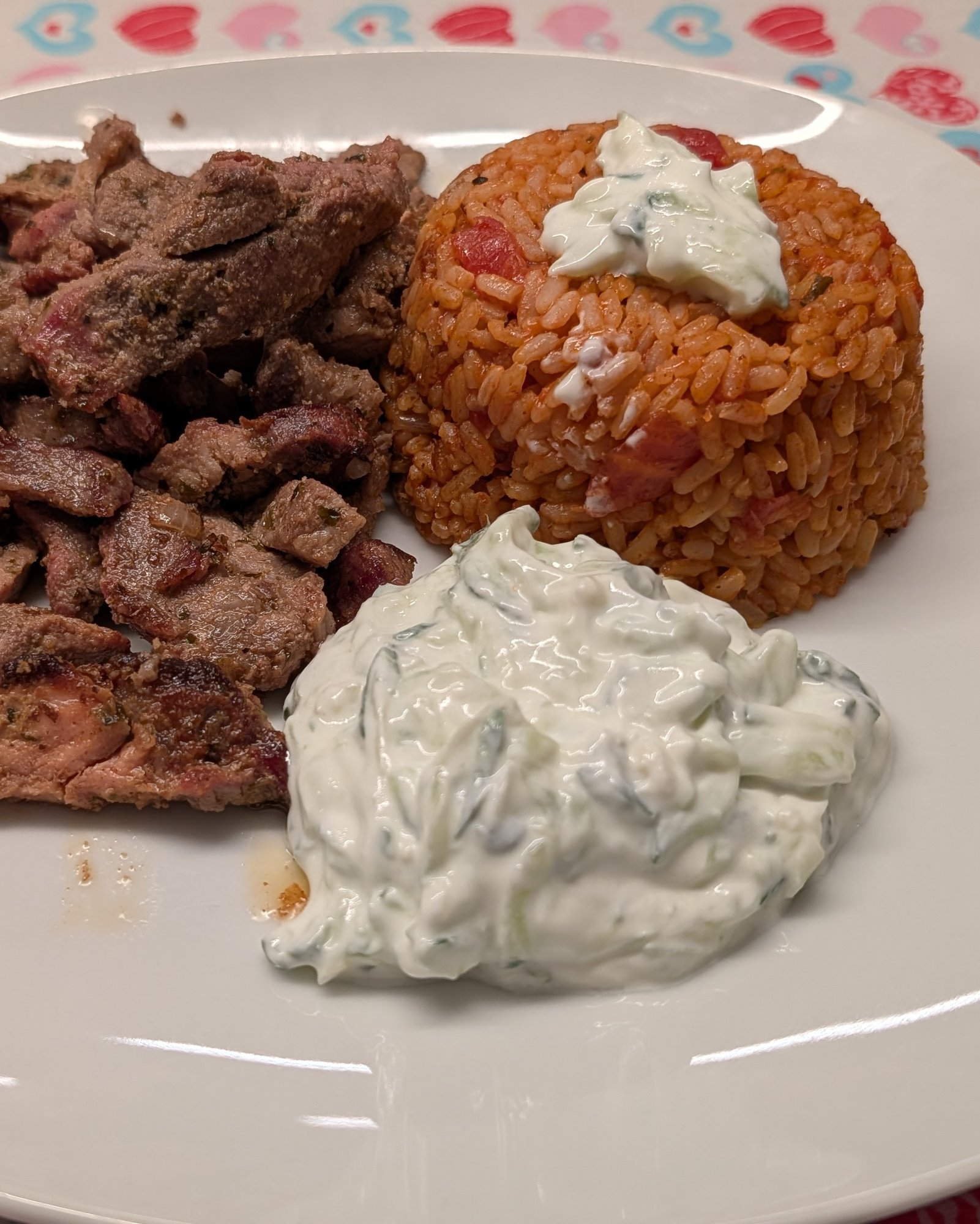 Assiette blanche carrée avec des morceaux de gyros saisis, un dôme de riz à la tomate surmonté de tzatziki et une portion de tzatziki au concombre