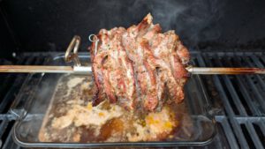 Tranches de porc marinées serrées sur une broche rôtissoire dans un gril à gaz, avec un plat en verre rempli de jus de cuisson en dessous