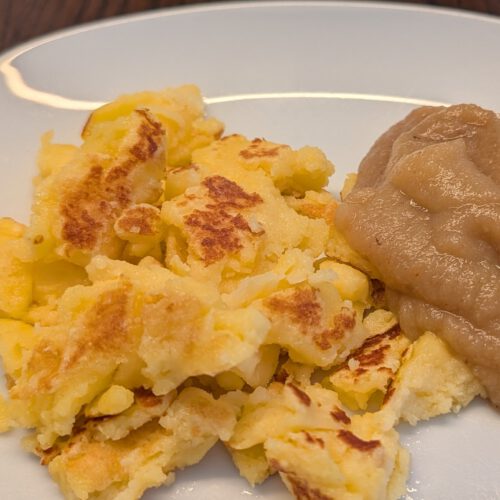 Morceaux dorés de Kaiserschmarrn déchiré et une louche de compote de pommes sur une assiette blanche, posée sur une table en bois sombre
