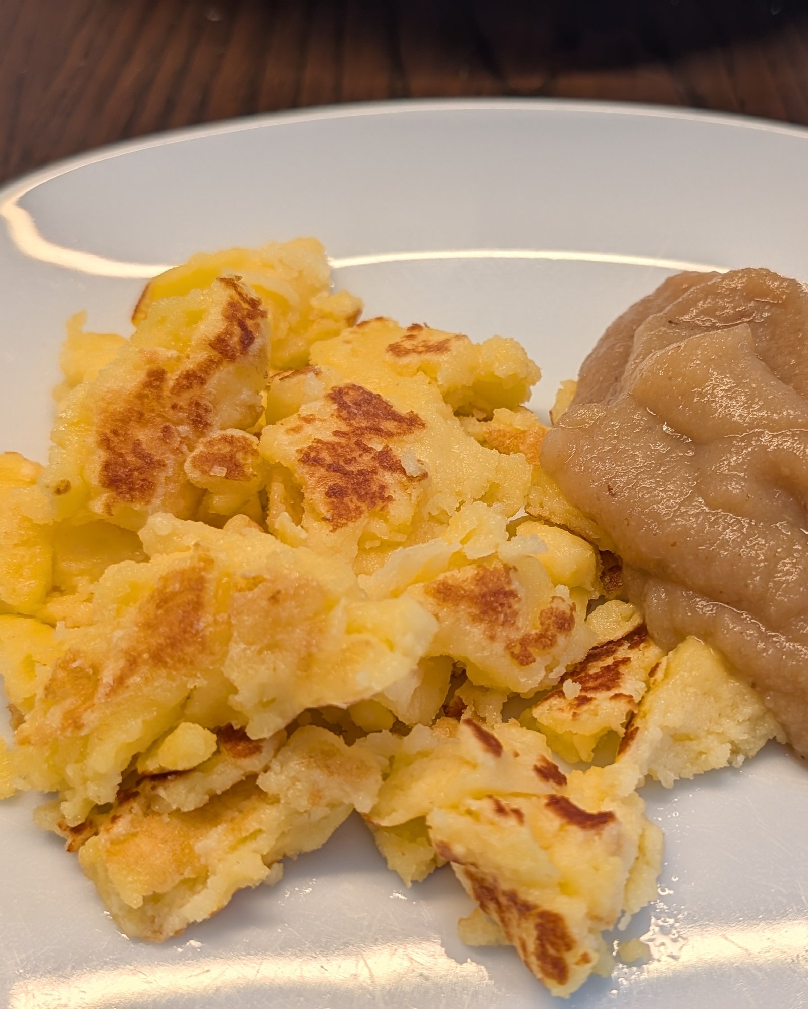 Morceaux dorés de Kaiserschmarrn déchiré et une louche de compote de pommes sur une assiette blanche, posée sur une table en bois sombre