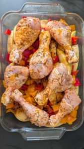 Pilons de poulet marinés et crus posés sur des rondelles de pommes de terre et des dés de poivron rouge et orange dans un plat en verre rectangulaire