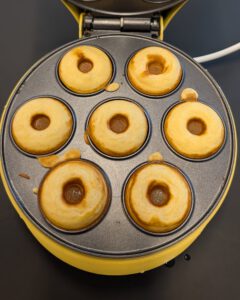 Sept mini donuts dorés fraîchement cuits dans les empreintes ouvertes d'une machine à donuts électrique jaune, vus du dessus