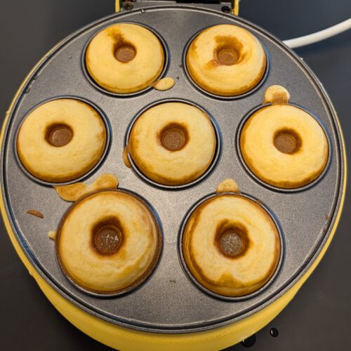 Sept mini donuts dorés fraîchement cuits dans les empreintes ouvertes d'une machine à donuts électrique jaune, vus du dessus