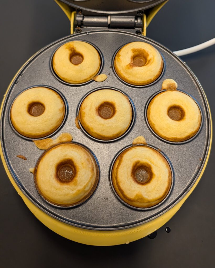 Sept mini donuts dorés fraîchement cuits dans les empreintes ouvertes d'une machine à donuts électrique jaune, vus du dessus