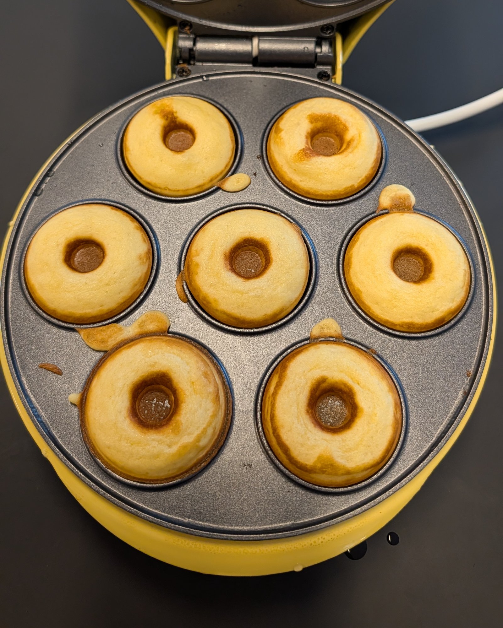 Sept mini donuts dorés fraîchement cuits dans les empreintes ouvertes d'une machine à donuts électrique jaune, vus du dessus