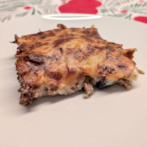Part de moussaka dans une assiette blanche, dessus doré moucheté de brun ; la viande hachée et des couches crémeuses pâles se voient sur la coupe