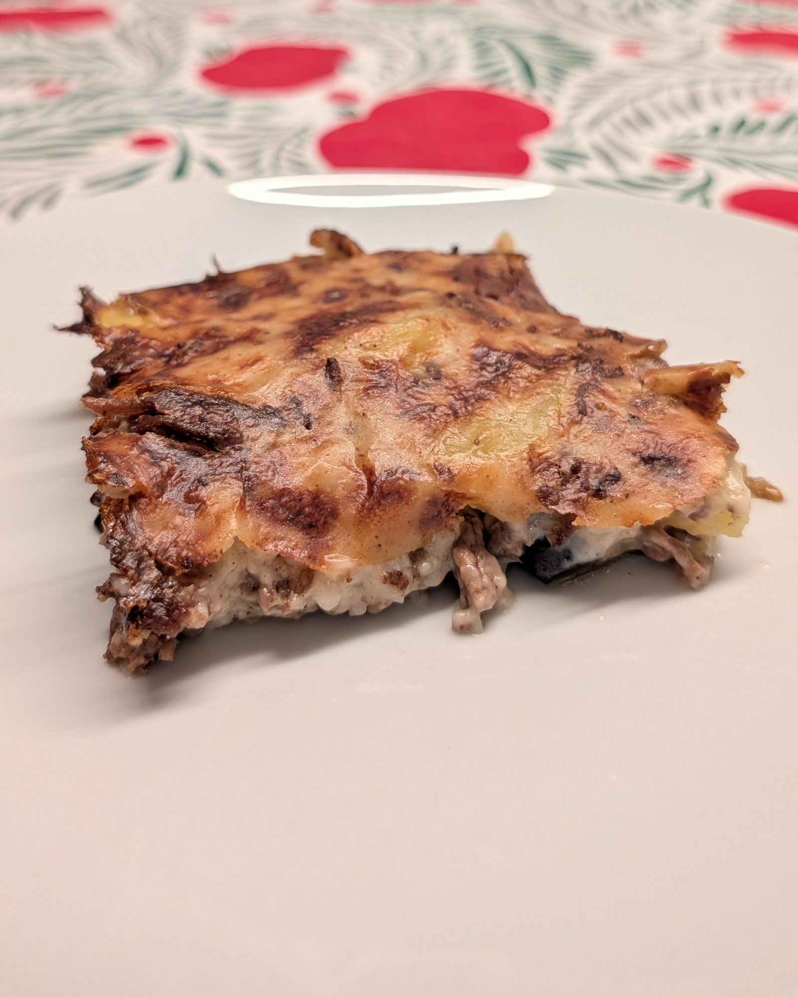 Part de moussaka dans une assiette blanche, dessus doré moucheté de brun ; la viande hachée et des couches crémeuses pâles se voient sur la coupe