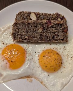 Deux tranches empilées de pain aux graines avec des fruits à coque au-dessus de deux œufs au plat poivrés sur une assiette blanche