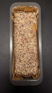 Pâte à pain aux graines tassée à plat dans un moule à cake garni de papier sulfurisé, vue du dessus, prête pour le repos au réfrigérateur