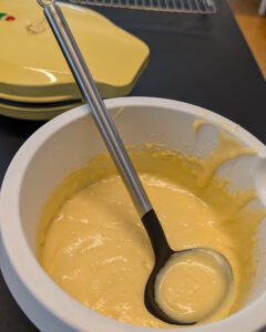 Pâte à donuts jaune et lisse dans un saladier blanc avec une louche, à côté d'une machine à donuts jaune fermée