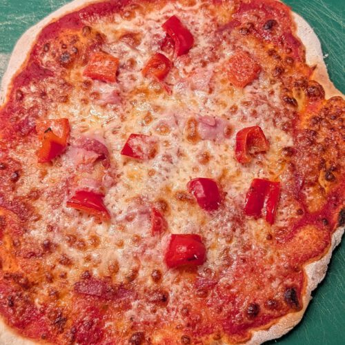 Pizza sans gluten cuite, avec sauce tomate, mozzarella fondue, morceaux de jambon et dés de poivron rouge sur une planche verte