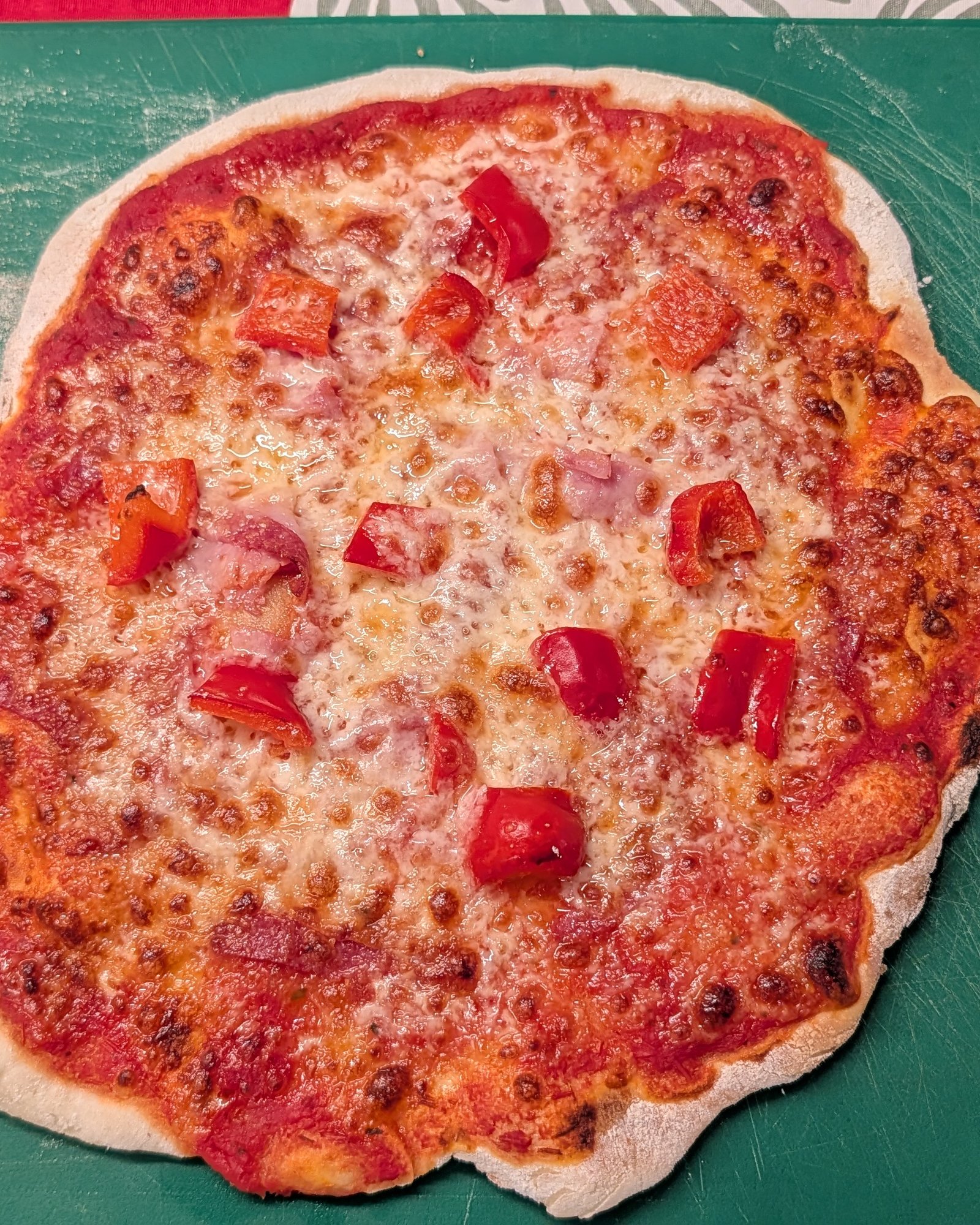 Pizza sans gluten cuite, avec sauce tomate, mozzarella fondue, morceaux de jambon et dés de poivron rouge sur une planche verte