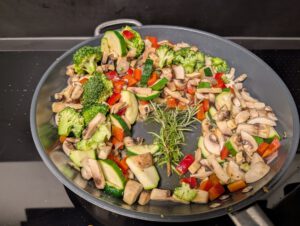 Brocoli, champignons, courgette et poivron rouge disposés sur le bord d'une poêle, avec des brins de romarin au milieu