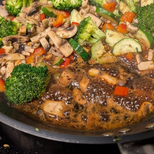 Poêle remplie de bouquets de brocoli, de lamelles de champignons, de rondelles de courgette et de morceaux de poivron rouge, avec un sirop balsamique sombre qui bouillonne au fond