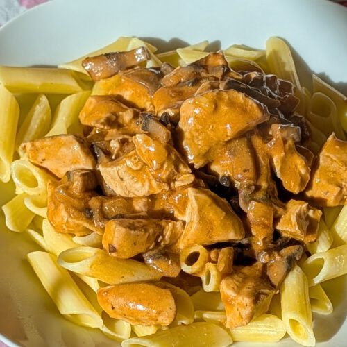 Poulet stroganoff crémeux aux champignons sur des penne sans gluten, dans une assiette blanche sur une nappe à cœurs, en plein soleil