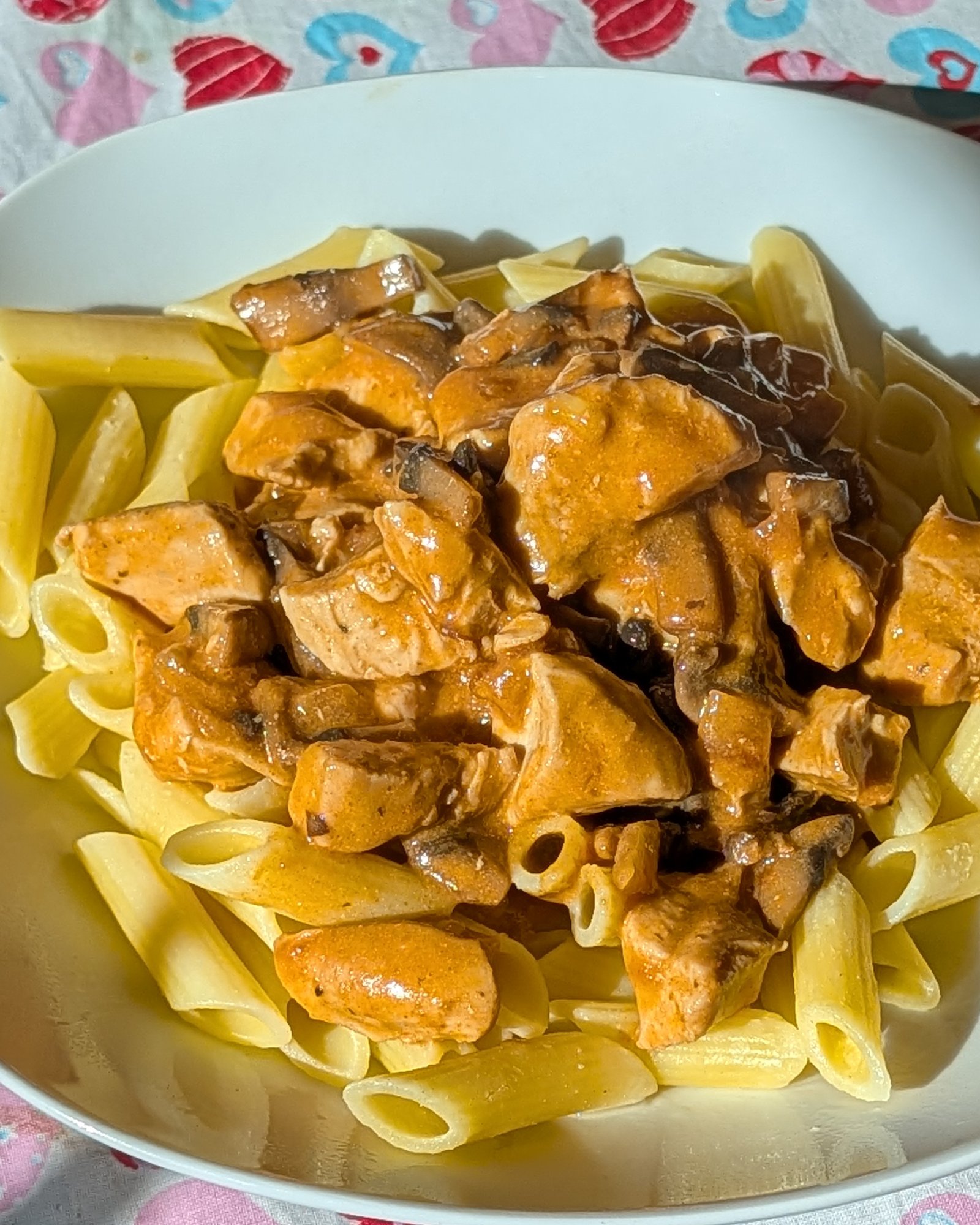 Poulet stroganoff crémeux aux champignons sur des penne sans gluten, dans une assiette blanche sur une nappe à cœurs, en plein soleil