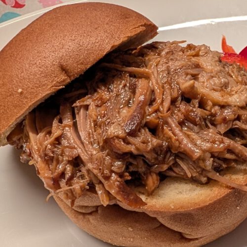 Pulled pork dans un pain à burger sans gluten, avec des lamelles de poivron rouge à côté, sur une assiette blanche devant une nappe à motif de cœurs