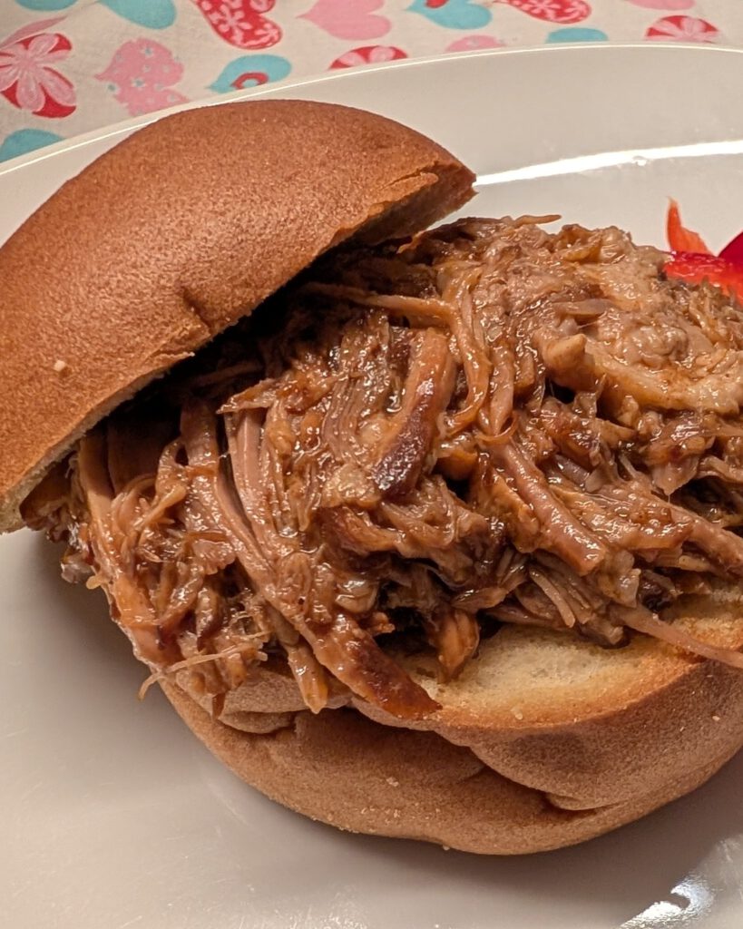 Pulled pork dans un pain à burger sans gluten, avec des lamelles de poivron rouge à côté, sur une assiette blanche devant une nappe à motif de cœurs