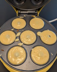 Pâte à donuts versée dans les sept empreintes d'une machine à donuts jaune ouverte, avec quelques gouttes sur l'assiette
