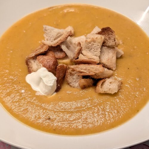 Soupe crémeuse orange dans une assiette creuse blanche et carrée, garnie de croûtons dorés et d'une cuillerée de crème aigre, sur une nappe à motif de cœurs