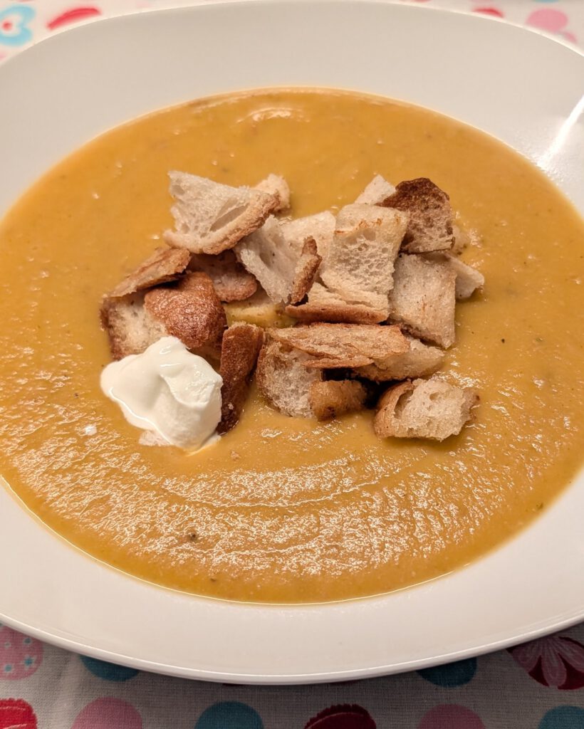 Soupe crémeuse orange dans une assiette creuse blanche et carrée, garnie de croûtons dorés et d'une cuillerée de crème aigre, sur une nappe à motif de cœurs